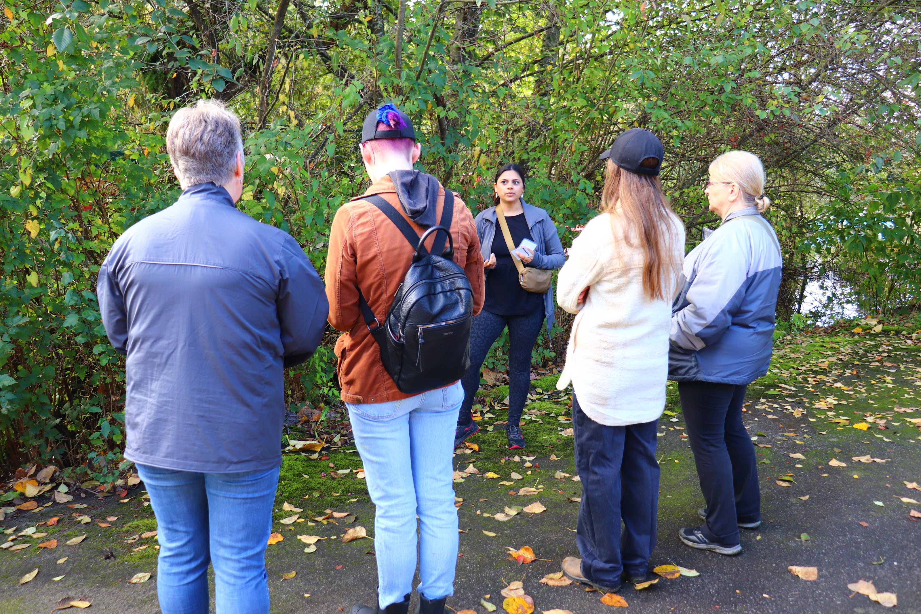Group of people standing outdoors in a park with trees and fallen leaves.