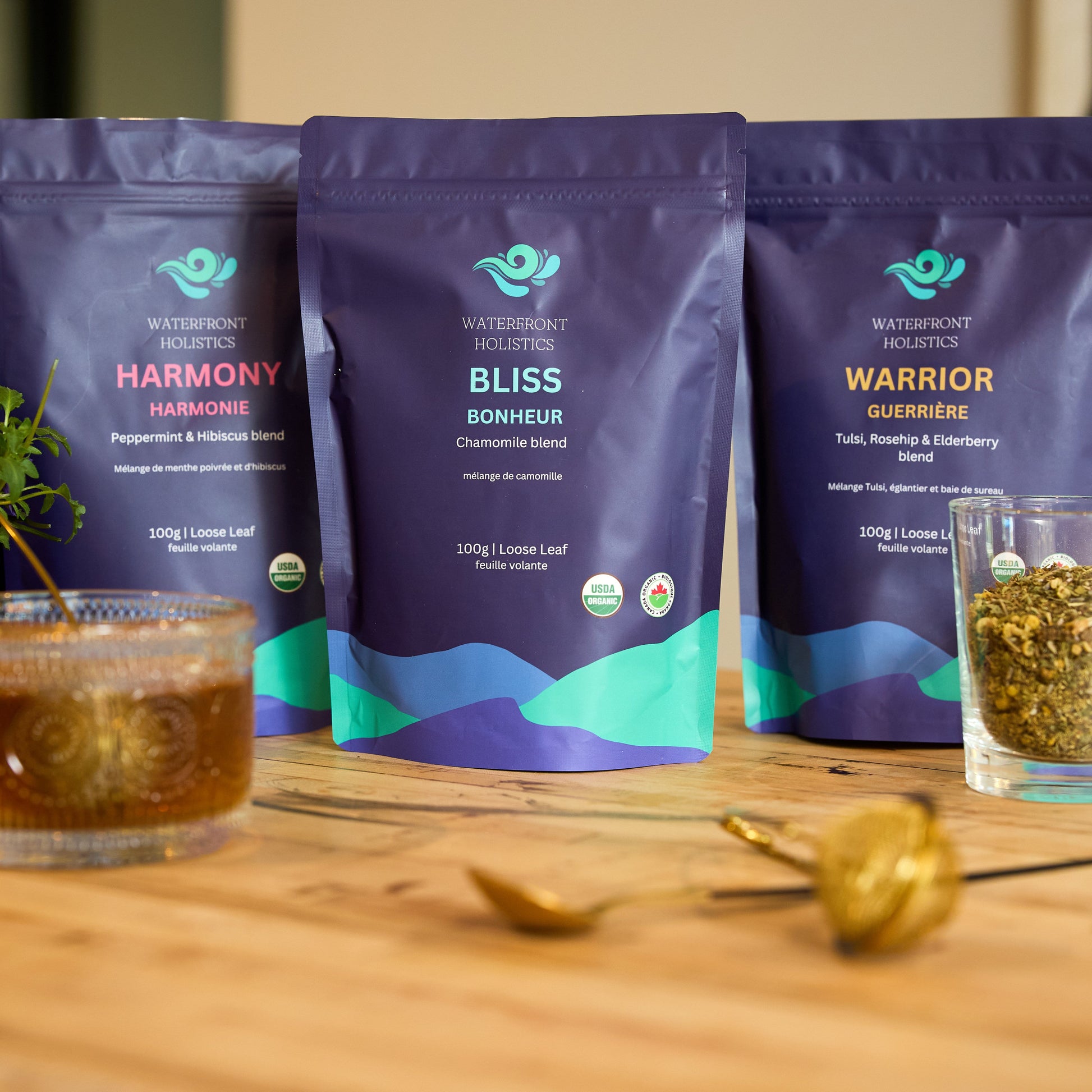 Three packages of Waterfront Holistics tea on a wooden surface with glasses of tea and flowers.
